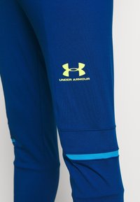 Modré sportovní kalhoty vyrobené z hladkého materiálu mají žluté logo Under Armour a modrý vodorovný pruh na dolní části nohy.