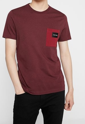 Bordowa koszulka z krótkim rękawem z ciemnoczerwonym kieszonką na piersi, na której znajduje się mała czarno-biała naszywka z logo Calvin Klein.