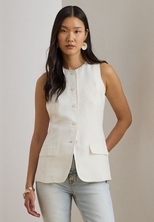 Frau mit langen dunklen Haaren trägt ein ärmelloses weißes, geknöpftes Weste, hellblaue Jeans, Perlenarmband und große runde Ohrringe, steht drinnen.