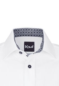 Weißes Hemd mit einem farbigen kragenfutter in Marineblau, ausgestattet mit schwarzen Knöpfen und einem Etikett mit dem "Kauf"-Logo. Glatte Stofftextur.