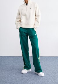 Gräddzip-svetter med dragkedja och gröna träningbyxor med gula detaljer och sidostrecken. Vita sneakers fulländar outfiten.