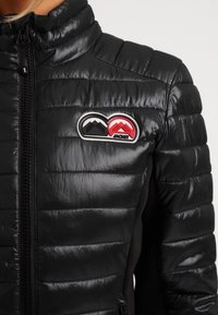 Veste matelassée noire avec zip devant et un patch sur la poitrine montrant des logos de montagnes noirs et rouges avec le mot "JACKIT."