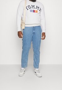 Felpa grigia chiaro con scritta "TOMMY" e disegni della bandiera, jeans in denim blu e scarpe da ginnastica bianche con dettagli navy. Outfit casual per l'uso quotidiano.