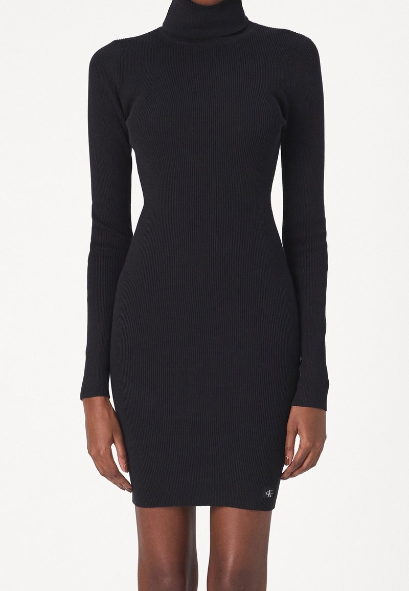 Robe noire ajustée en maille côtelée avec manches longues et col roulé, portée par une personne debout sur un fond blanc uni.