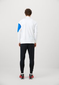 Hvid atletisk jakke med blå accent på venstre skulder, parret med sorte tapered bukser og rødt fodtøj. Glat stoftekstur.