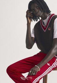 Άτομο με dreadlocks που φοράει καφέ γιλέκο, λευκό μπλουζάκι και κόκκινο αθλητικό παντελόνι, κρατώντας γυαλιά ενώ κάθεται σε ξύλινο σκαμπό.