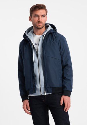 Giacca blu navy con cappuccio, dotata di zip frontale, tasche laterali e polsini a coste. Indossata sopra un felpa azzurro chiaro e una maglietta bianca.