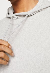 Sudadera gris claro hecha de un tejido suave y texturizado, con cierre de cordón, cuello ajustado y detalles de costura visibles.