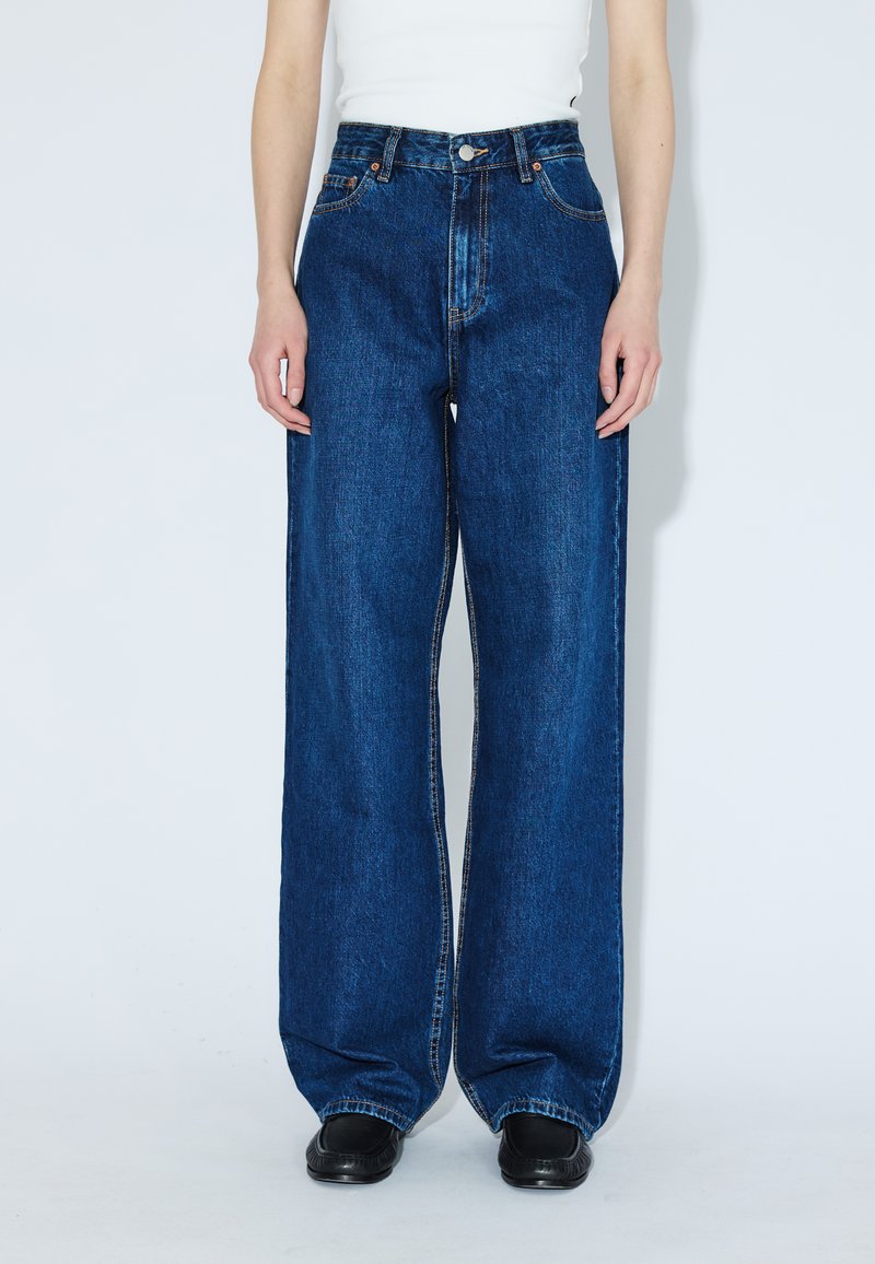 Person iført blå jeans med højtaljede vide ben, hvid ærmeløs top og sorte sko, der står mod en ensfarvet hvid baggrund.