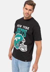 Černé bavlněné tričko s velkým grafickým potiskem helmy New York Jets v zelené a bílé barvě, s textem, krátkými rukávy a volným střihem.