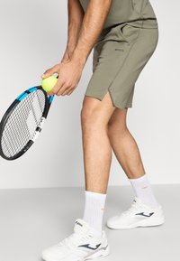 Λαδί αθλητικό σορτς με πλευρικές σχισμές, σε συνδυασμό με λευκά αθλητικά παπούτσια και ριμπ λευκές κάλτσες, κρατώντας κίτρινη μπάλα του τένις και ρακέτα.