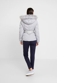 Femme de dos portant une doudoune gris clair avec capuche bordée de fourrure, pantalon bleu marine et baskets bleu clair sur fond blanc.