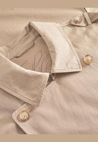 Camisa de manga corta beige con cuello, dos botones redondos y detalles interiores cosidos. Textura suave con un diseño simple y limpio.