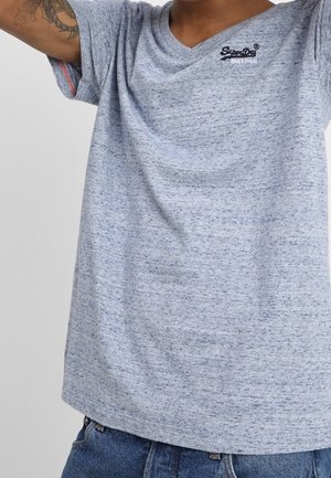 Osoba ubrana w jasnoszary melanżowy t-shirt z małym logo "SuperDry" na klatce piersiowej oraz niebieskie dżinsy, unosząca obie ręce.