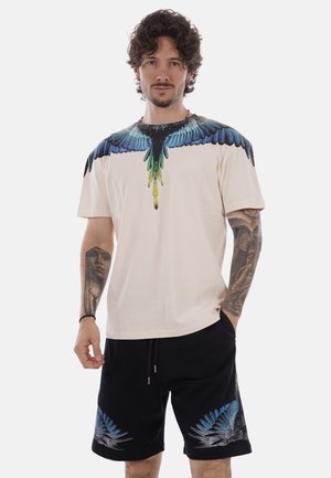 Mann mit lockigem Haar und Tätowierungen auf beiden Armen trägt ein beiges T-Shirt mit blau-grünem Vogelflügel-Print und schwarze Shorts mit blauen Flügelmotiven.