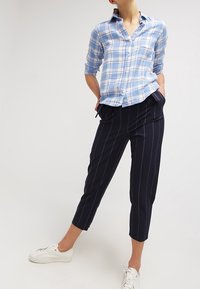 Světle modrá kostkovaná košile s dlouhými rukávy, spárovaná s tmavě modrými pruhovanými kalhotami se zkrácenou délkou. Oblečení doplňují bílé tenisky.