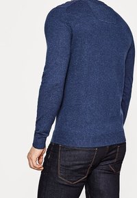 Mörkblå tröja i mjukt stickat material med rund halsringning och ribbade ärmslut och nederkant, som bärs över mörka jeans, sedd bakifrån.