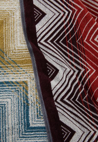 Gewoven deken met zigzagpatronen in mosterd, blauw, bordeauxrood en roomkleur. Zachte textuur met een gelaagd ontwerp en contrasterende randen.