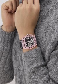 Muñeca con un reloj Guess de cuadrante negro y rosa, con dos esferas, sobre un fondo de suéter gris de punto.