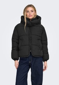 Musta toppatakki, jossa korkea kaulus, tikattu kuvio ja etuvetoketjusuljenta. Takissa on joustavat hihansuut ja lyhyt mitoitus.