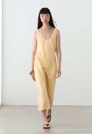 Femme marchant à l'intérieur portant une combinaison beige sans manches, des sandales noires, et tenant des bracelets en bois à son poignet gauche.
