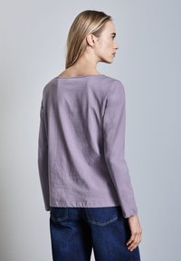 Langärmliges lila Baumwollhemd mit weitem Ausschnitt und geradem Saum, kombiniert mit dunkelblauen Jeans. Weiche Textur, minimalistisches Design.