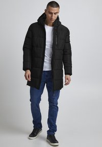 Sort pufferjakke med hætte, der har lodrette syninger, lynlås foran og justerbar forneden. Bæret over en hvid skjorte og blå jeans.
