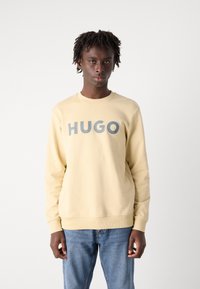 Lys gul sweatshirt med rund hals og ribbestrikkede mansjetter, med en grå "HUGO" logo på tvers av brystet, sammen med blå jeans.