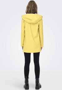 Gul regnjakke med hætte, lange ærmer og midt-lår længde. Parret med sorte leggings og sorte ankelstøvler. Glat tekstur.