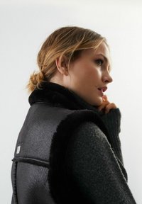 Czarna kamizelka o teksturowanym wykończeniu i miękkim kołnierzu, noszona na ciemnym, dzianinowym swetrze. Posiada małe logo z tyłu.