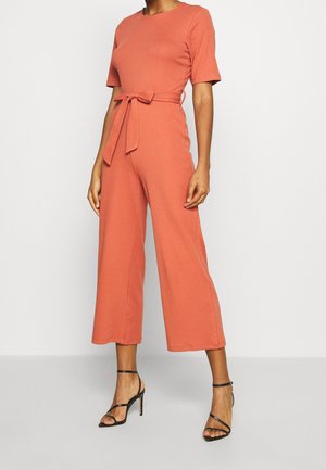 Ribbet jumpsuit i brændt orange med korte ærmer, bindebånd i taljen og bukser med vide ben. Kombineret med sorte sandaler med stropper.