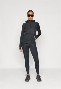Zwarte sportjack met een rits aan de voorkant en meshdetails, gecombineerd met zwarte leggings en gelijnde hardloopschoenen. Zonnebril op.