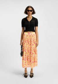 Schwarzes, geripptes Kurzarmoberteil mit Reißverschluss, kombiniert mit einem langen, gefältelten Rock mit einem rosa und gelben Blumenmuster. Schwarze Handtasche und Schuhe.