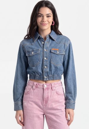 Giovane donna sorride indossando una giacca di jeans corta e jeans rosa a vita alta su uno sfondo semplice.