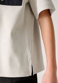 Helehall lühikeste varrukatega T-särk ümmarguse äärega, millel on külje lõhikud ja kontrastse musta plaastriga ülemisel seljal.