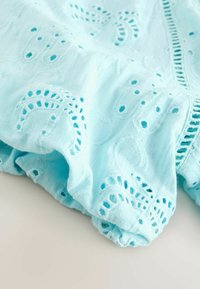 Világoskék anyag hímzett lyukacsos mintákkal és fodros, összegyűrt szegélyekkel, puha, texturált anyagon.