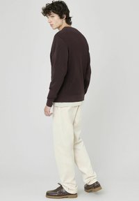 Chemise longue à manches ribbées marron avec un col rond, associée à un pantalon couleur crème et des chaussures à lacets marron, vue de dos.