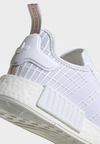 Zapatilla deportiva blanca con un upper de patrón en cuadrícula, suela texturizada y una lengüeta estriada en la parte trasera con letras rojas.