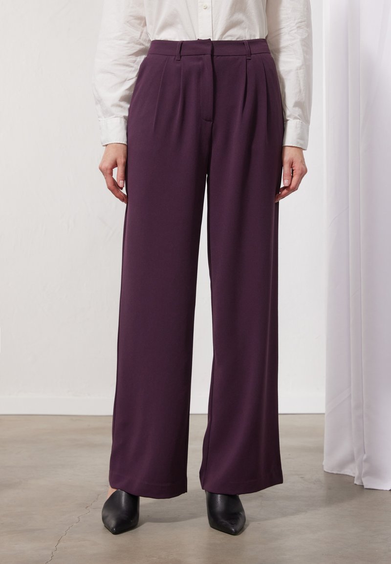 Wijde broek in diep paarse stof, met voorplooien en een op maat gemaakte tailleband, gecombineerd met scherpe zwarte hakken.