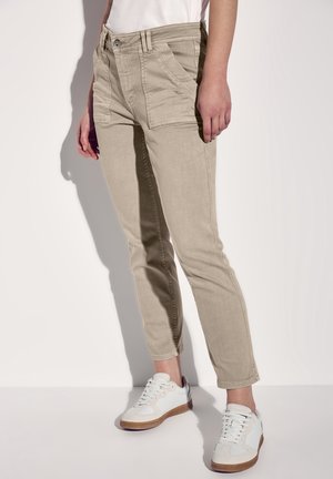 Personne portant un pantalon beige slim-fit avec poches plaquées devant et des baskets blanches, debout devant un fond clair.