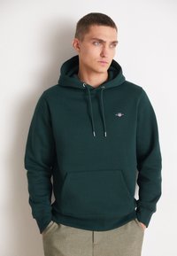 Dark green hoodie made from soft cotton, featuring a front pocket, drawstring hood, and a small logo in the chest area.