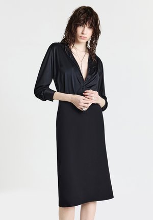 Abito nero con bottoni, realizzato in materiale liscio, con maniche lunghe, scollo a V e silhouette dritta che arriva fino a metà polpaccio.