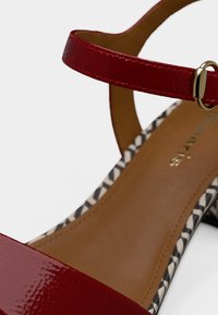 Κόκκινο σανδάλι από δέρμα με λεπτό λουράκι στον αστράγαλο και χρυσά μεταλλικά στοιχεία. Η εσωτερική σόλα έχει ένα ασπρόμαυρο γεωμετρικό σχέδιο.