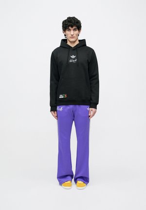 Sudadera con capucha negra con logo plateado y parche, combinada con pantalones morados con franjas laterales. Zapatos amarillos completan el conjunto.