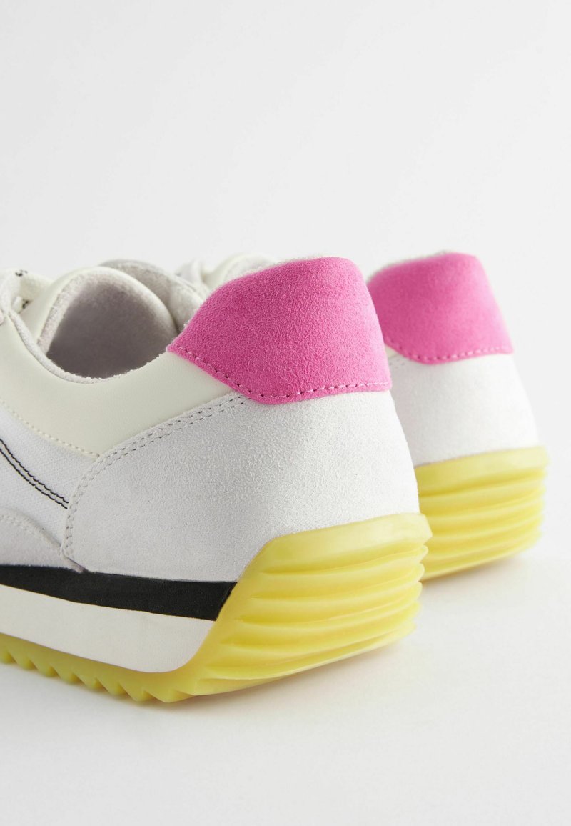 Weiße Sportschuhe mit rosa Wildlederakzenten an der Ferse, schwarzem Mittelsohlenstreifen und strukturiertem gelben Gummisohle. Mit Schnürverschluss und runder Form.