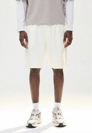 Mies, joka pukeutunut valkoisiin, pinnaltaan kuvioituihin polvipitkiin shortseihin, harmaisiin kerrospaitoihin, valkoisiin lyhyisiin sukkiin ja valkoisiin urheilutennareihin, seisoo yksinkertaisen taustan edessä.