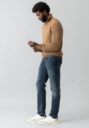 Brauner Rippstrickpullover mit einem Viertel-Reißverschluss, kombiniert mit dunkelblauen Jeans und weißen Sneakers mit grauen Details. Das Model steht im Profil.