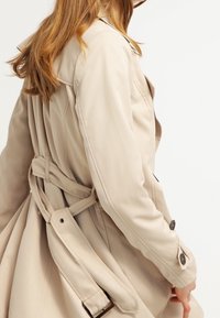 Beżowy płaszcz trencz o dopasowanym kroju, z paskiem wiązanym z tyłu oraz guzikami na mankietach. Gładka faktura i smukła sylwetka.