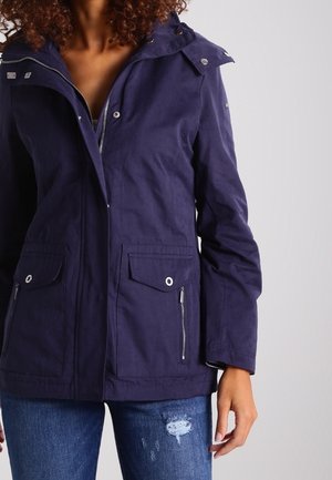 Vrouw draagt een marineblauw jack met rits- en knoopzakken, gecombineerd met blauwe gescheurde spijkerbroek, staand tegen een effen achtergrond.