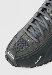 Γκρι αθλητικό παπούτσι με συνδυασμό λείων και υφασμάτινων υλικών, με πάνελ από διχτυωτό υλικό, χοντρά κορδόνια και μια σχέδιο ελαστική σόλα.